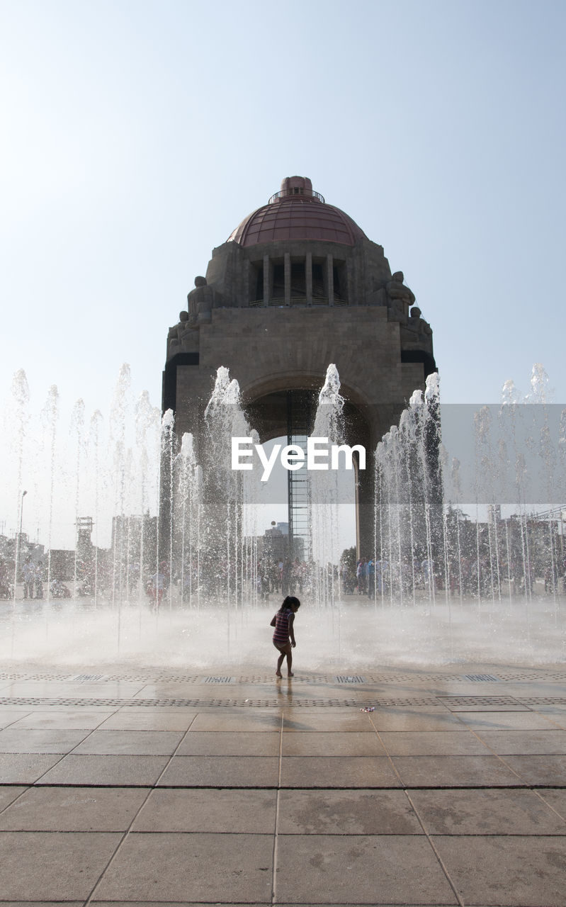 Girl standing by fountain in city against clear sky