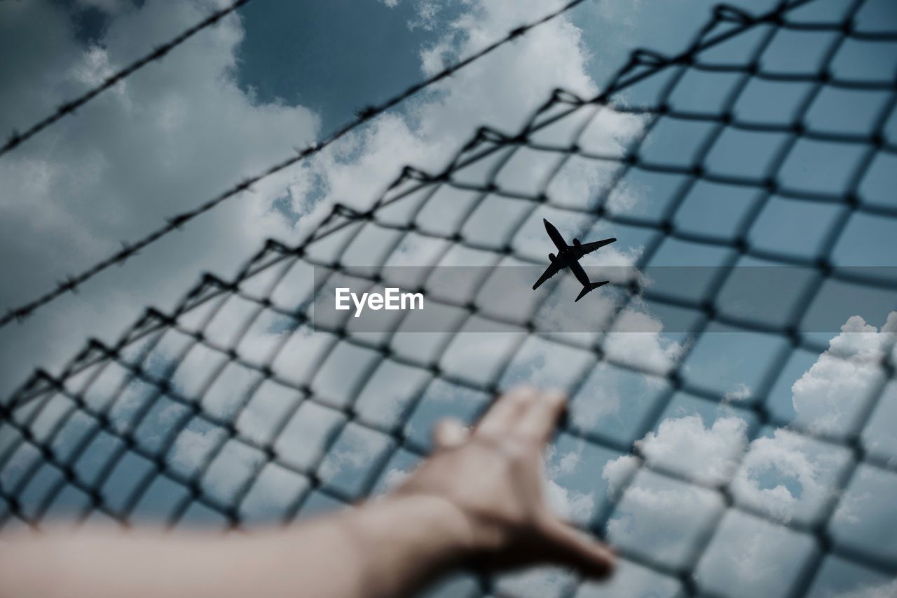 Cropped hand holding fence against airplane flying in sky