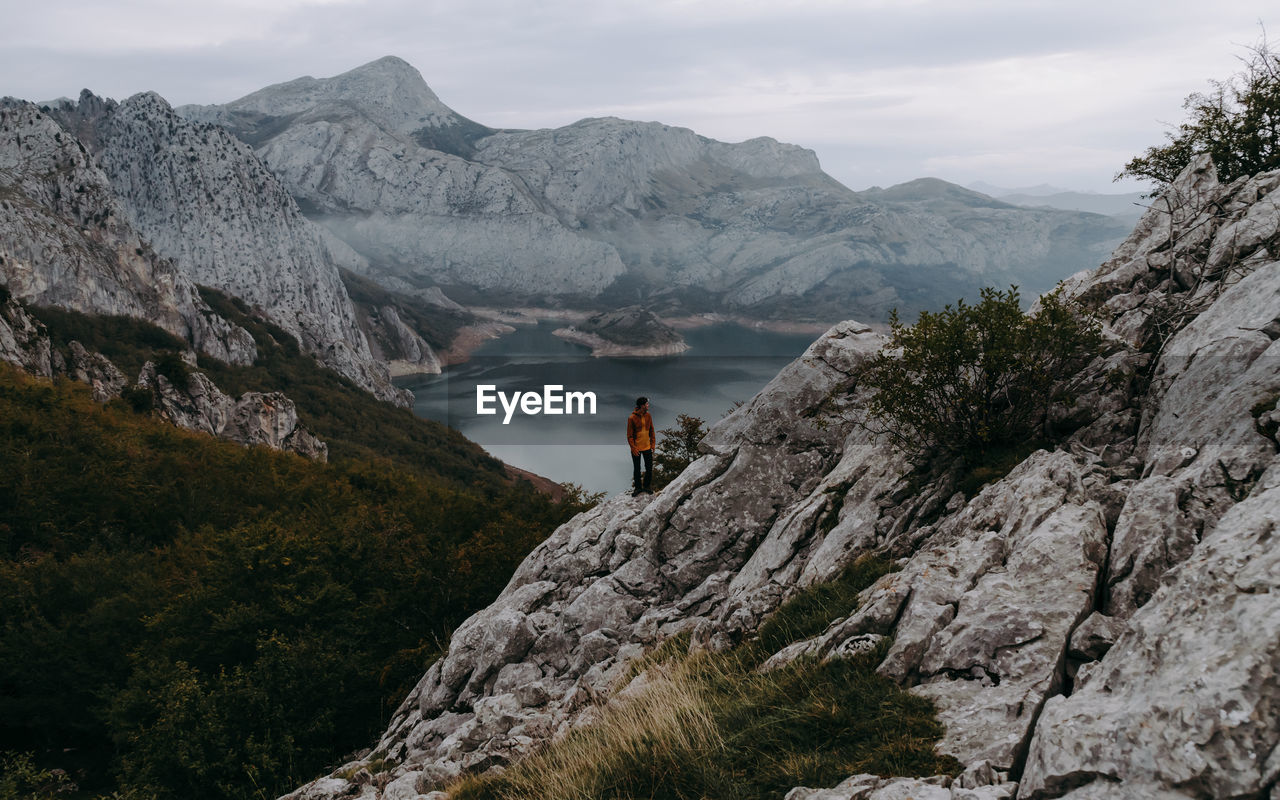 Man on rocks by mountains and lake agaisnt cloudy sky