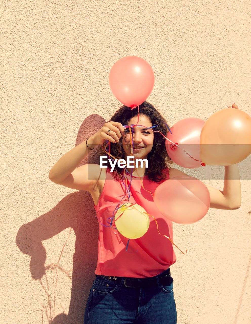 Smiling young woman holding balloons against wall during sunny day