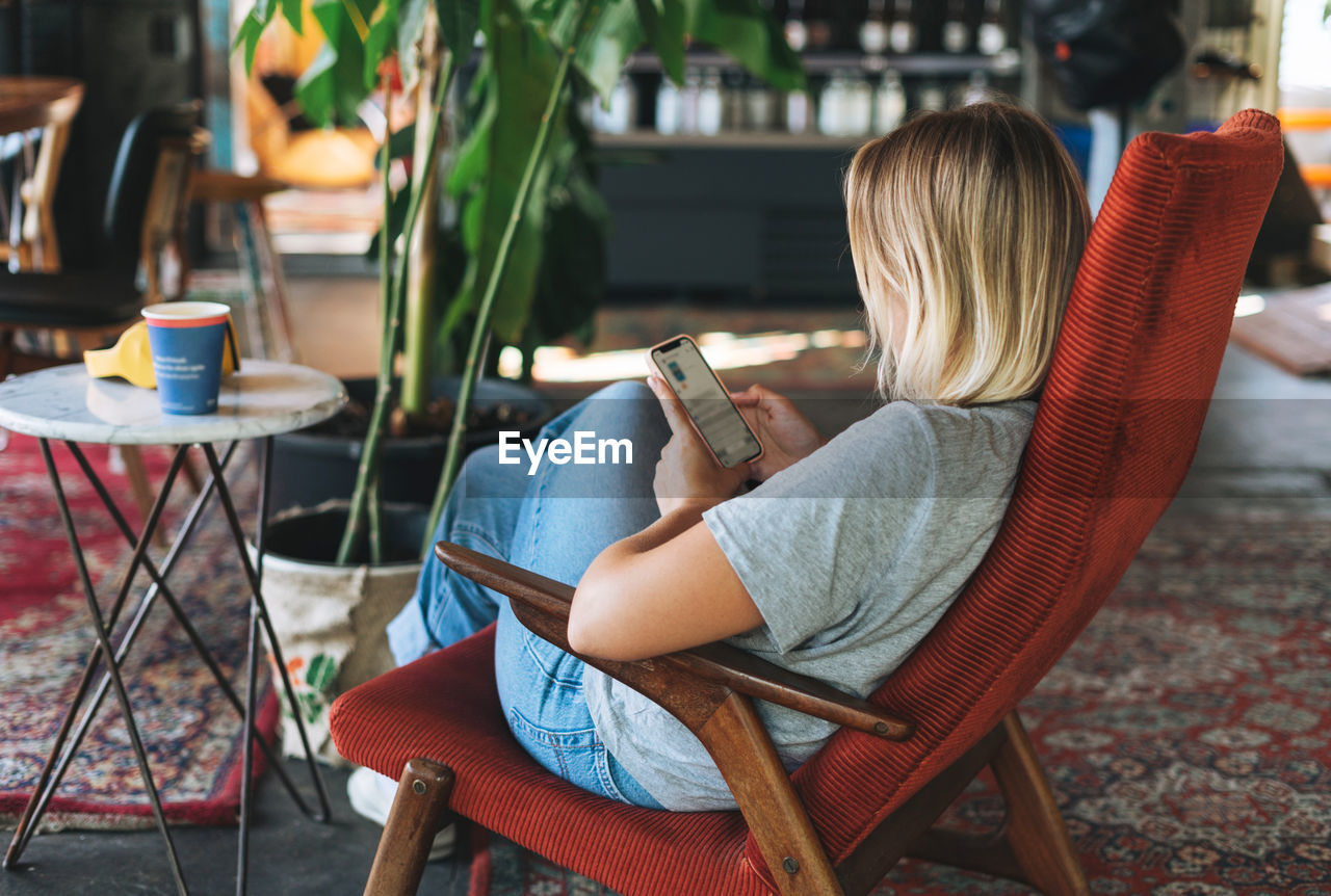 Young blonde woman in casual clothes using social media on mobile phone sitting on the chair at cafe