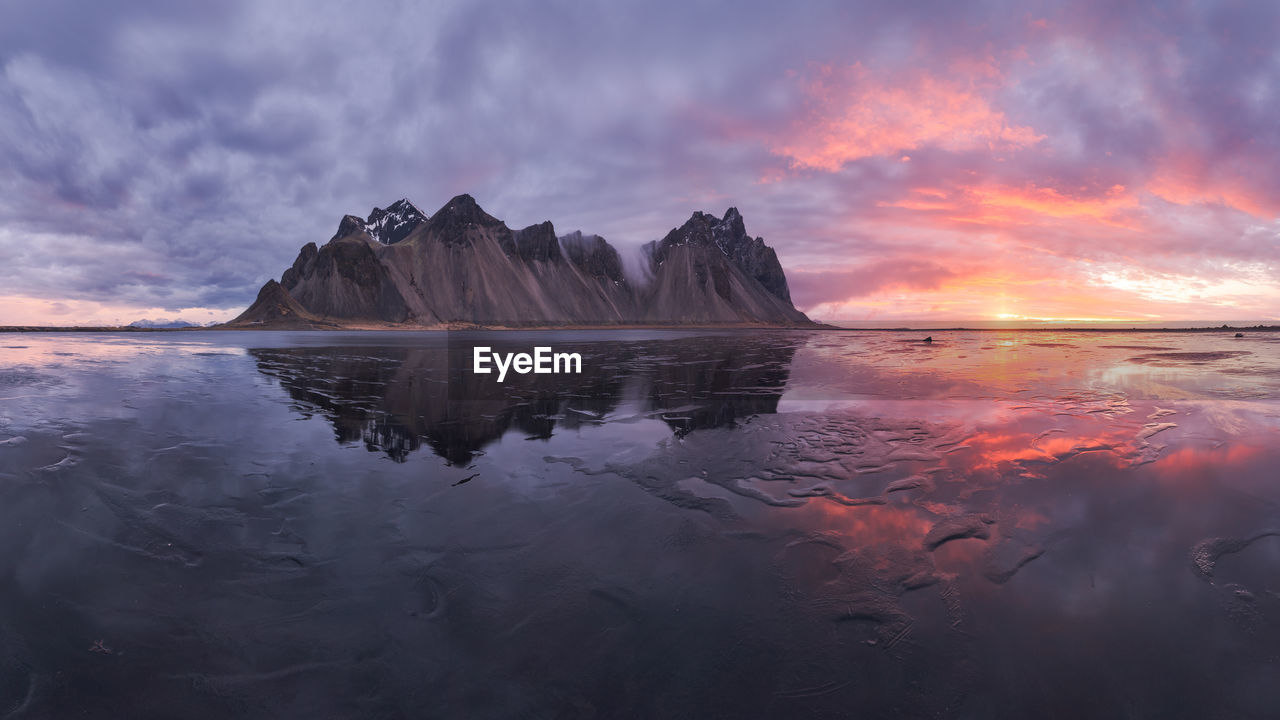 Spectacular nordic scenery of calm frozen lake near rocky vestrahorn mountain with snowy peaks during colorful sunset at stockness beach, iceland