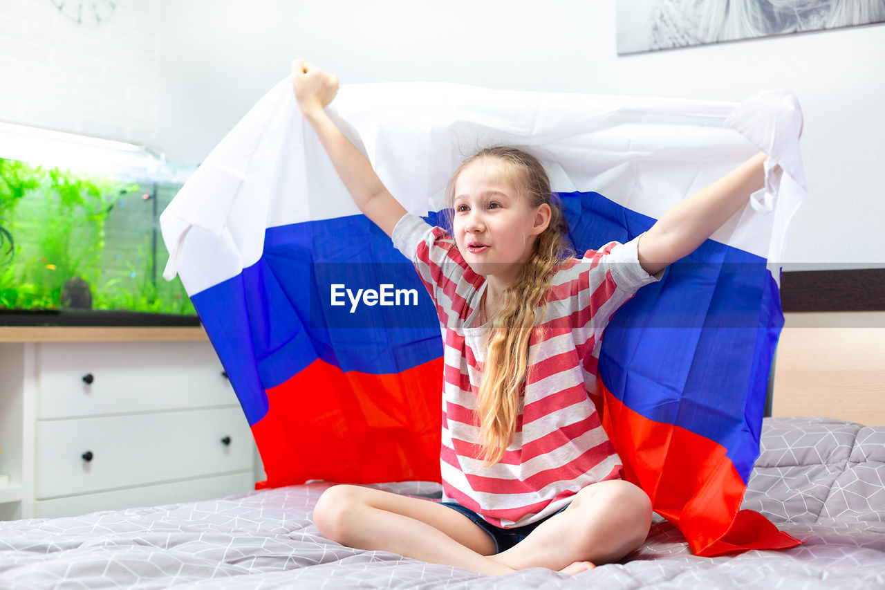 Happy beautiful caucasian girl with the russian flag in raised hands on the bed in front of the tv.