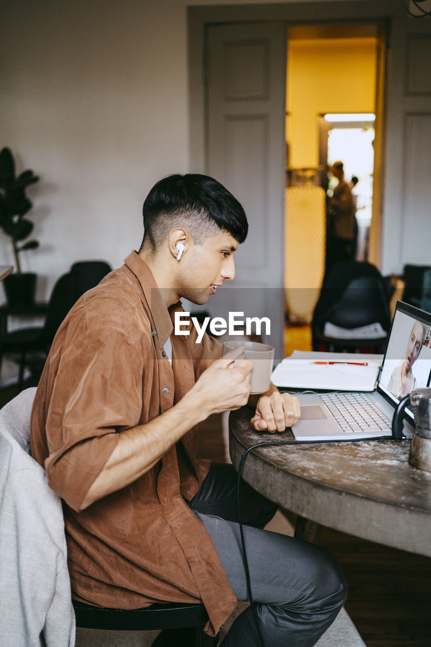 Side view of male freelancer having coffee while doing video call with colleague on laptop at home office