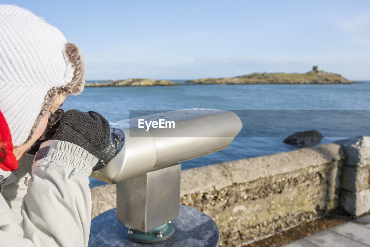 Side view of young woman looking through coin-operated binoculars while standing against sky