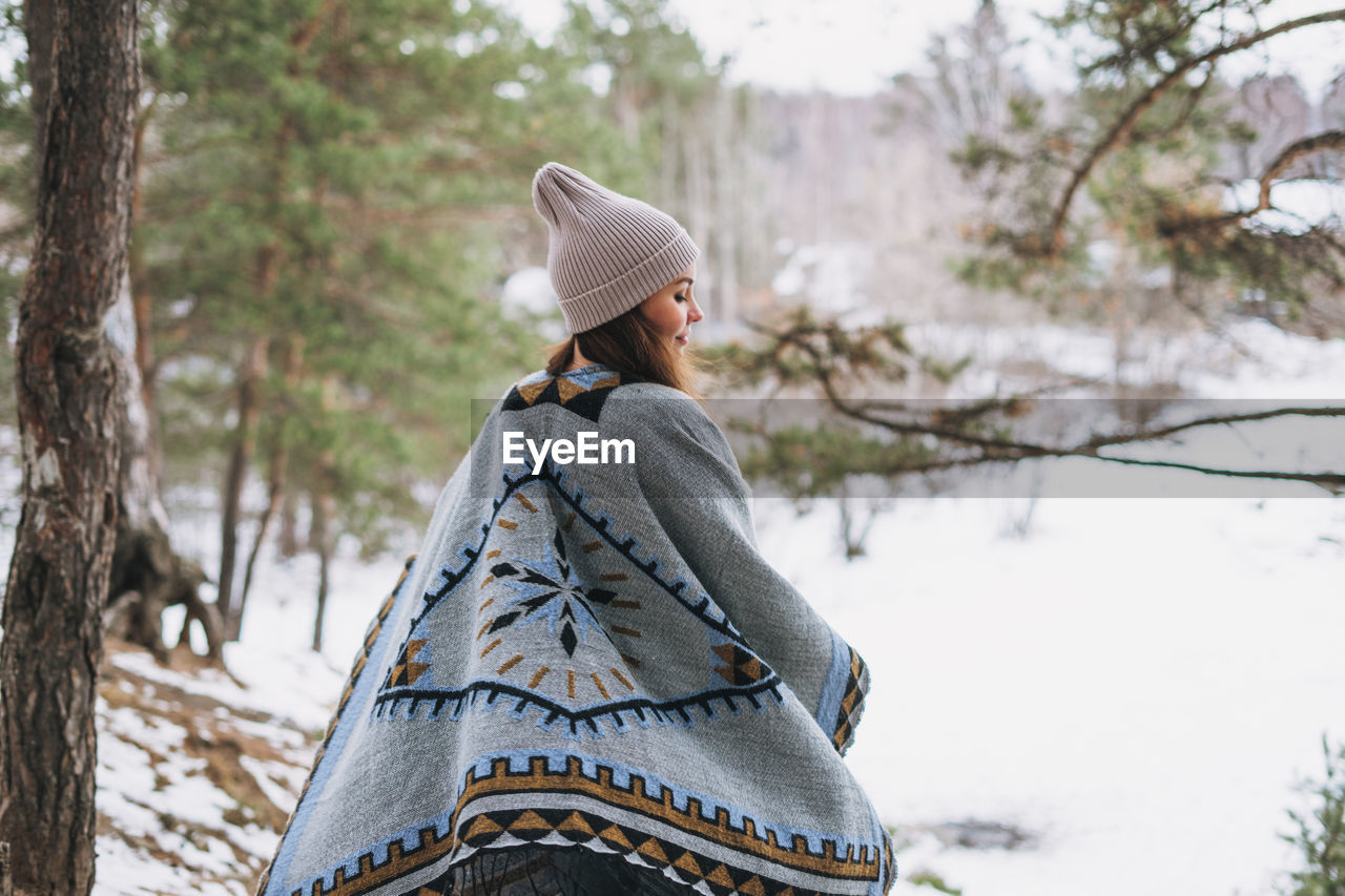 Young brunette beautiful woman in hat and grey poncho walking in the winter forest
