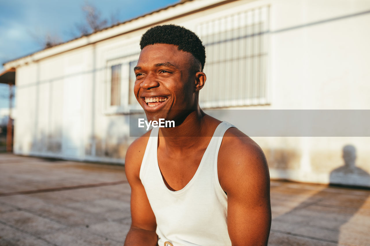 Happy african american male in undershirt with modern haircut looking forward against building in sunlight