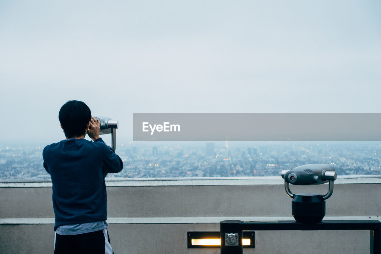 Rear view of man looking through binoculars against sky during foggy weather