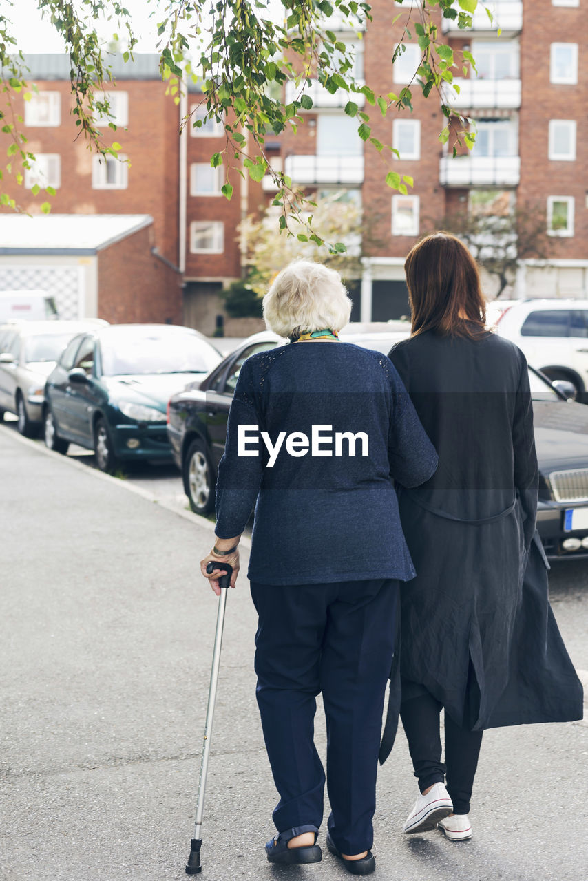 Rear view of elderly woman walking with granddaughter on sidewalk in city