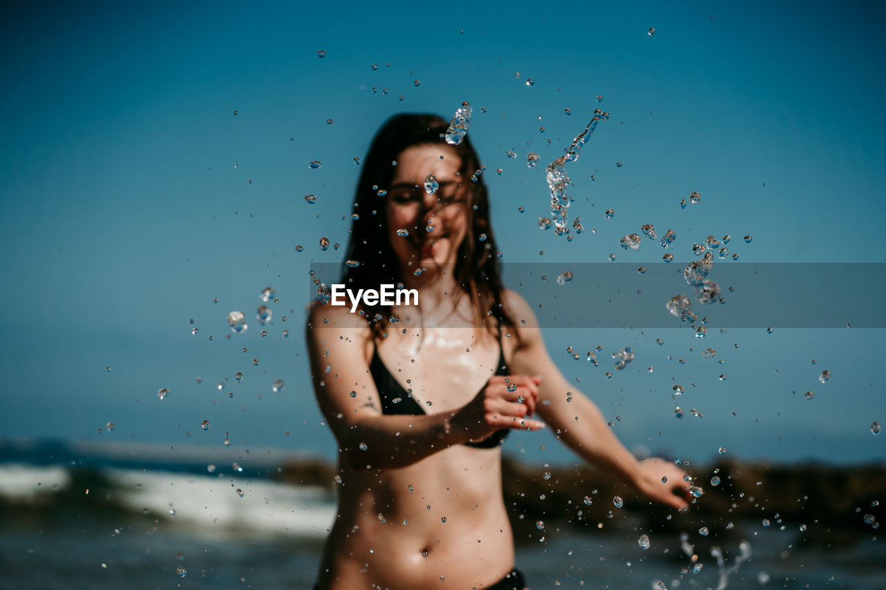 Cheerful female in bikini splashing clear water of sea while having fun in summer during holiday