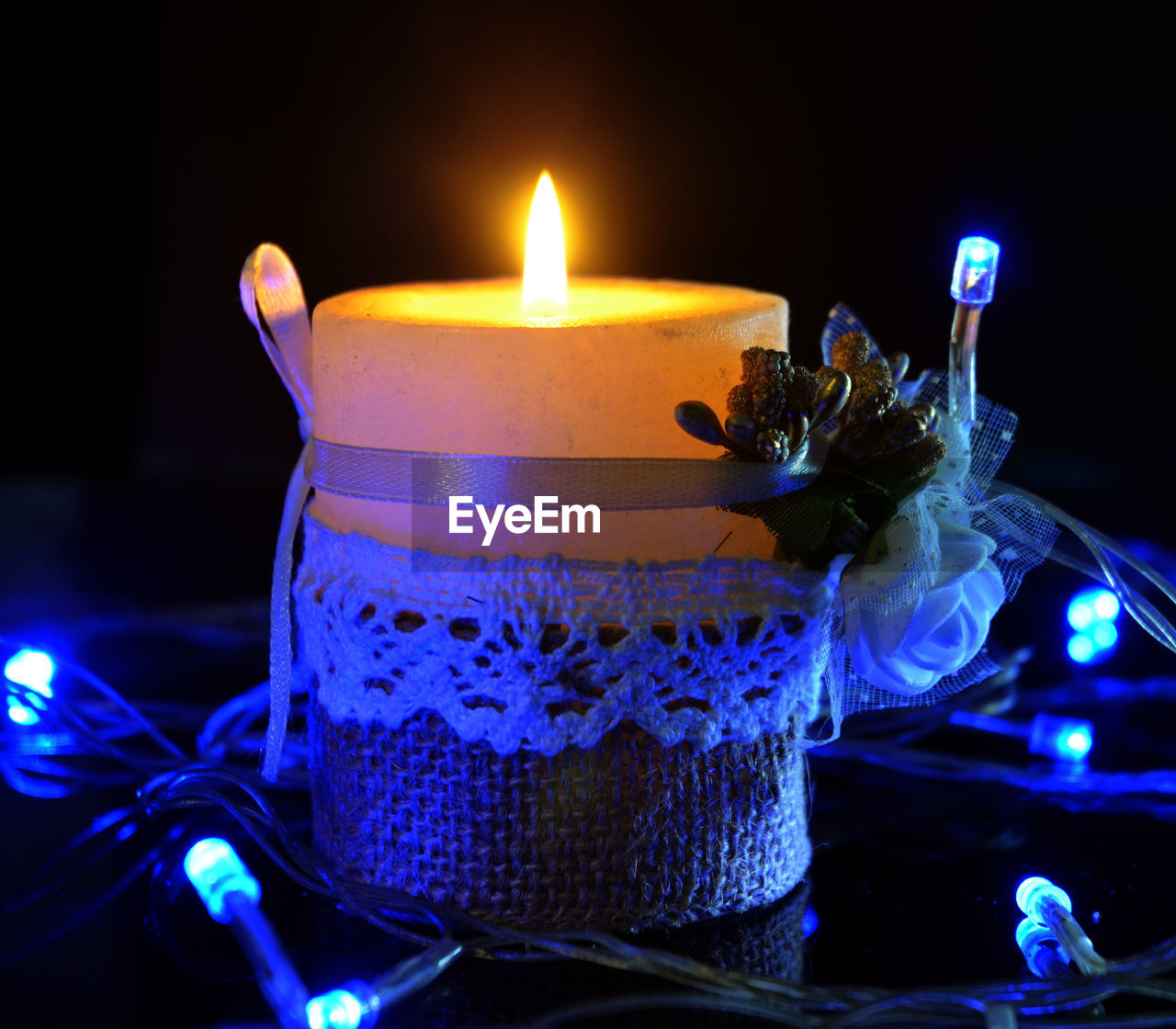 Close-up of lit candle with illuminated string lights on table against black background