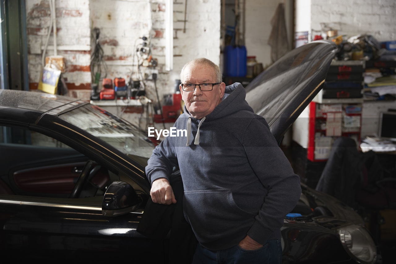 Confident male mechanic with hand in pocket leaning on car at workshop
