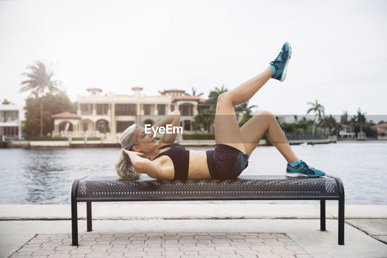 Side view of mature woman doing sit-ups on bench by river