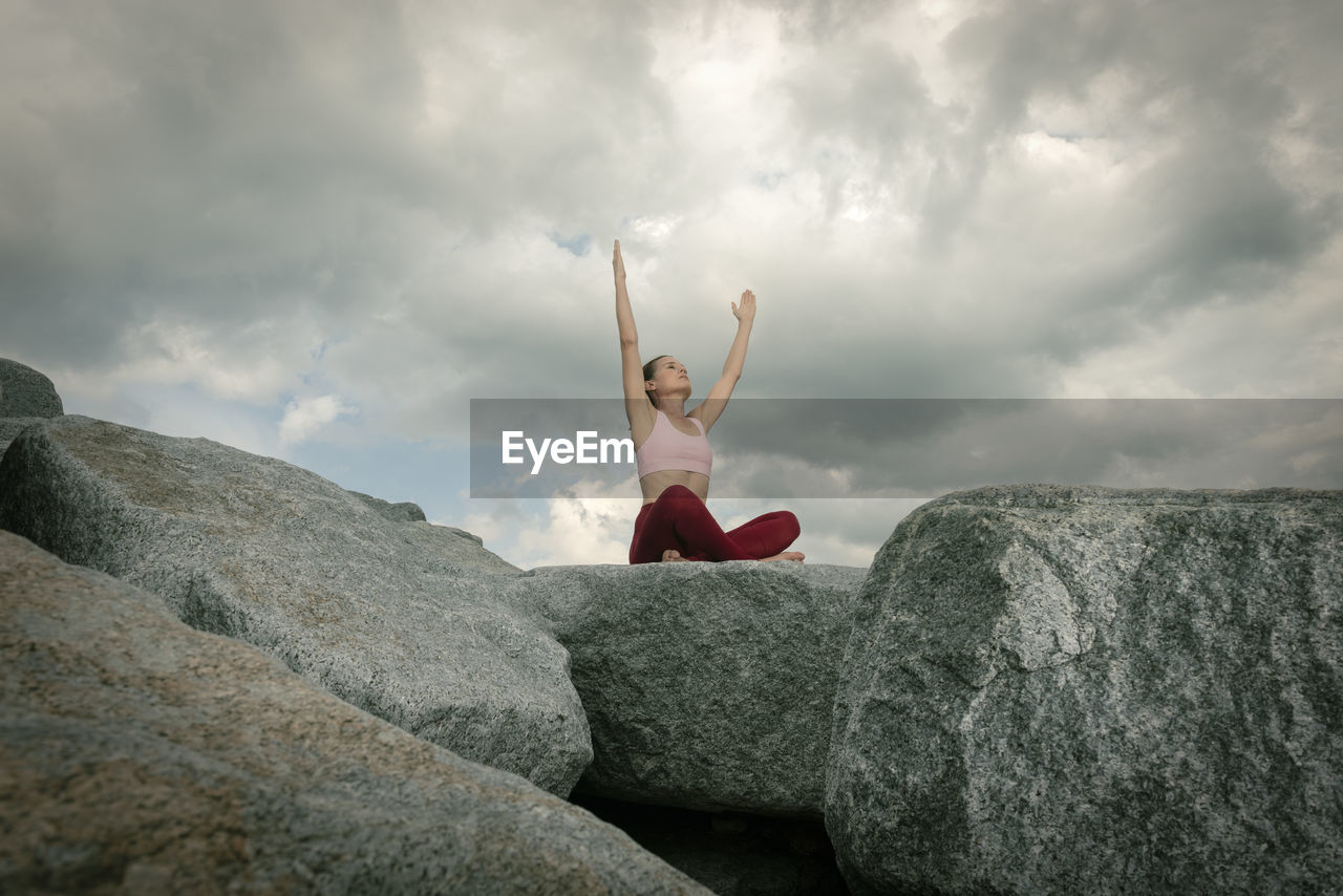 Sporty woman sitting crossed legged on rocks meditating and practicing yoga, dramatic sky.