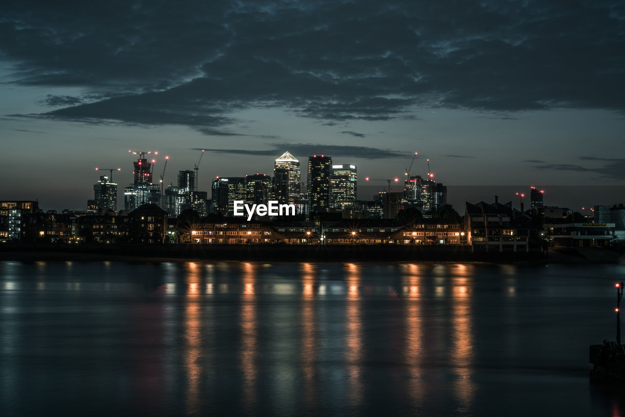 Illuminated buildings by river against sky at night