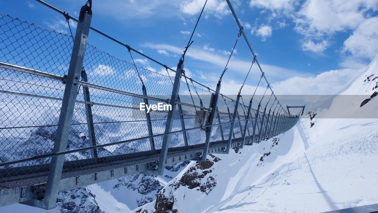 Footbridge on snowcapped mountain against cloudy sky