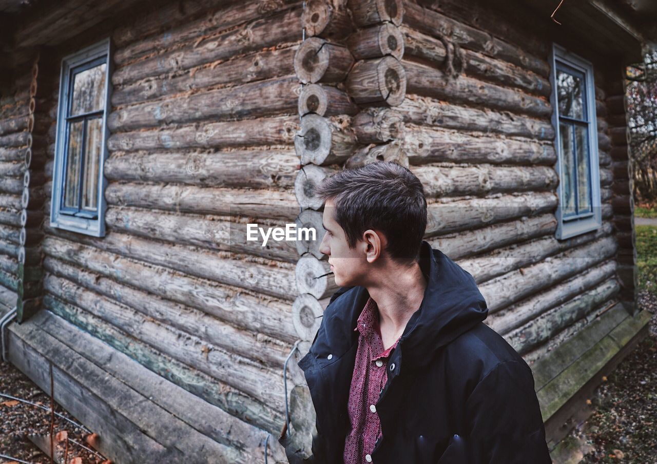 Man looking away while standing by log cabin