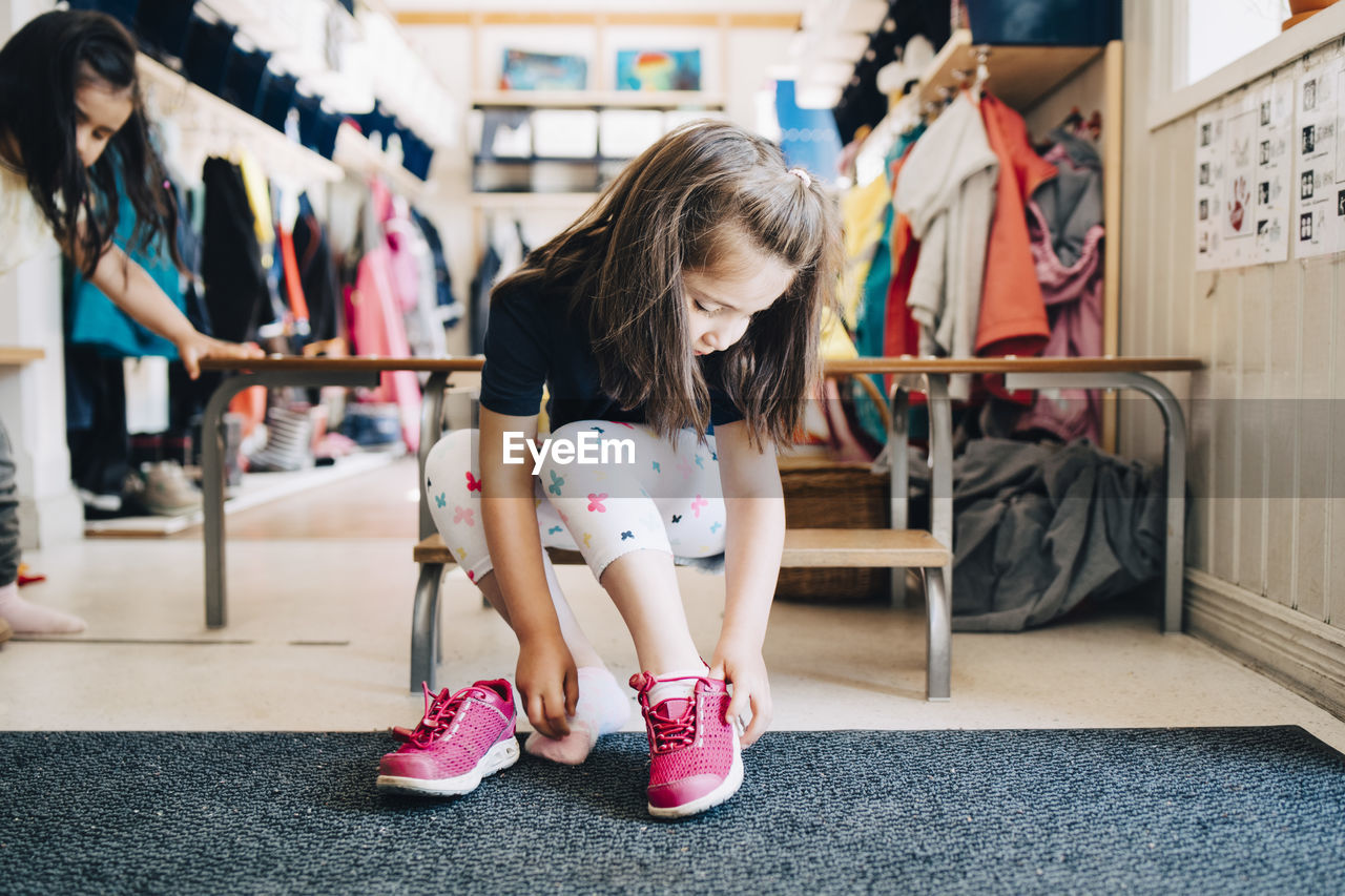 Full length of girl sitting on bench wearing shoes in cloakroom at preschool