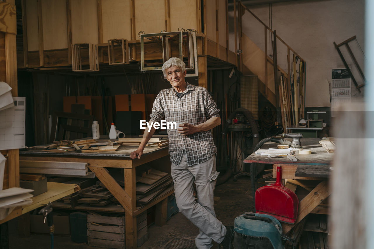 Smiling senior craftsman leaning on workbench at carpentry workshop