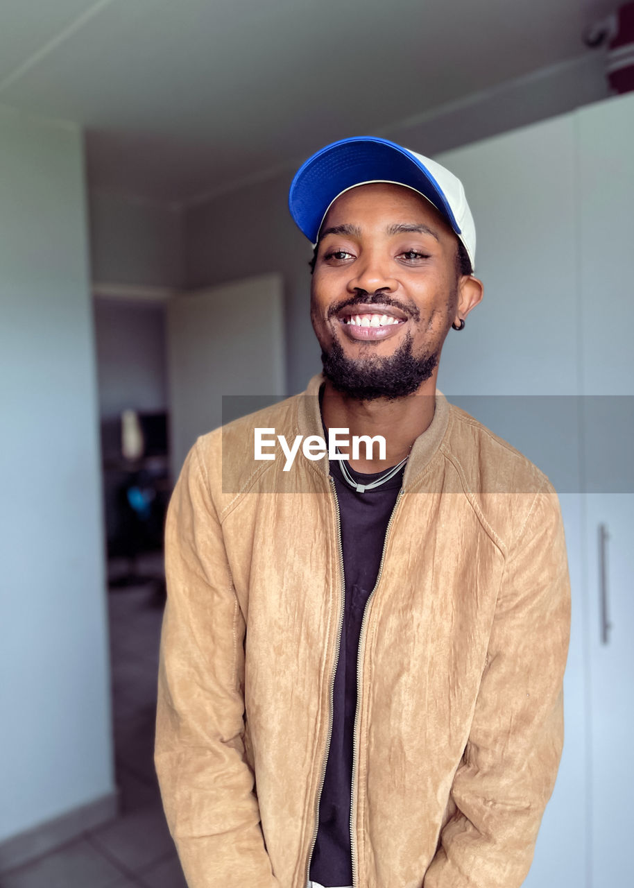 one person, adult, smiling, happiness, portrait, men, indoors, young adult, standing, beard, clothing, looking at camera, fashion, cheerful, facial hair, emotion, lifestyles, smile, teeth, waist up, hat, person, casual clothing, cool attitude, looking, front view, business, architecture, occupation, human face, cap, stubble, jacket, music