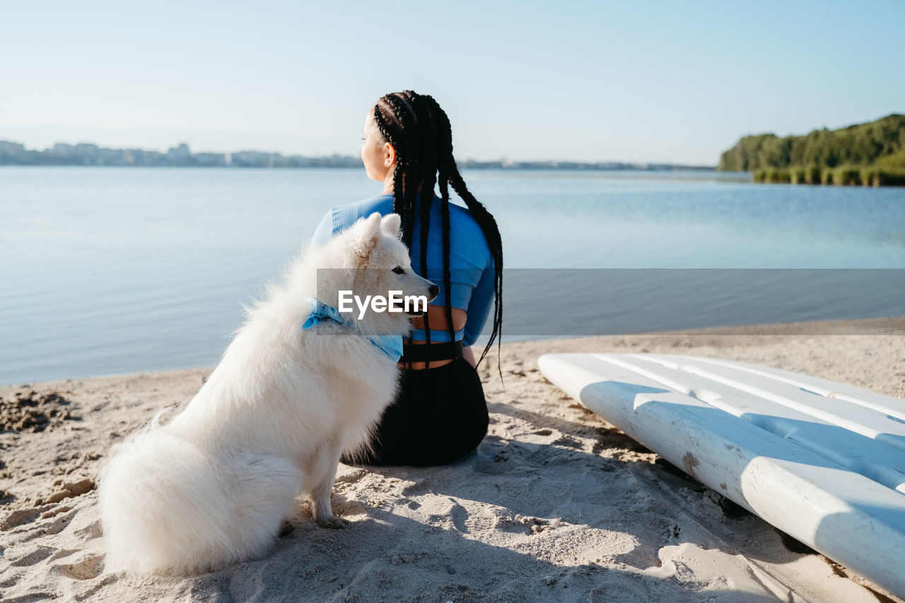 Woman with locs sitting on the beach of city lake with her best friend, dog breed japanese spitz