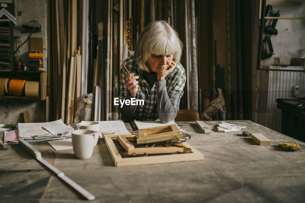 Thoughtful senior female carpenter holding pencil while leaning on workbench