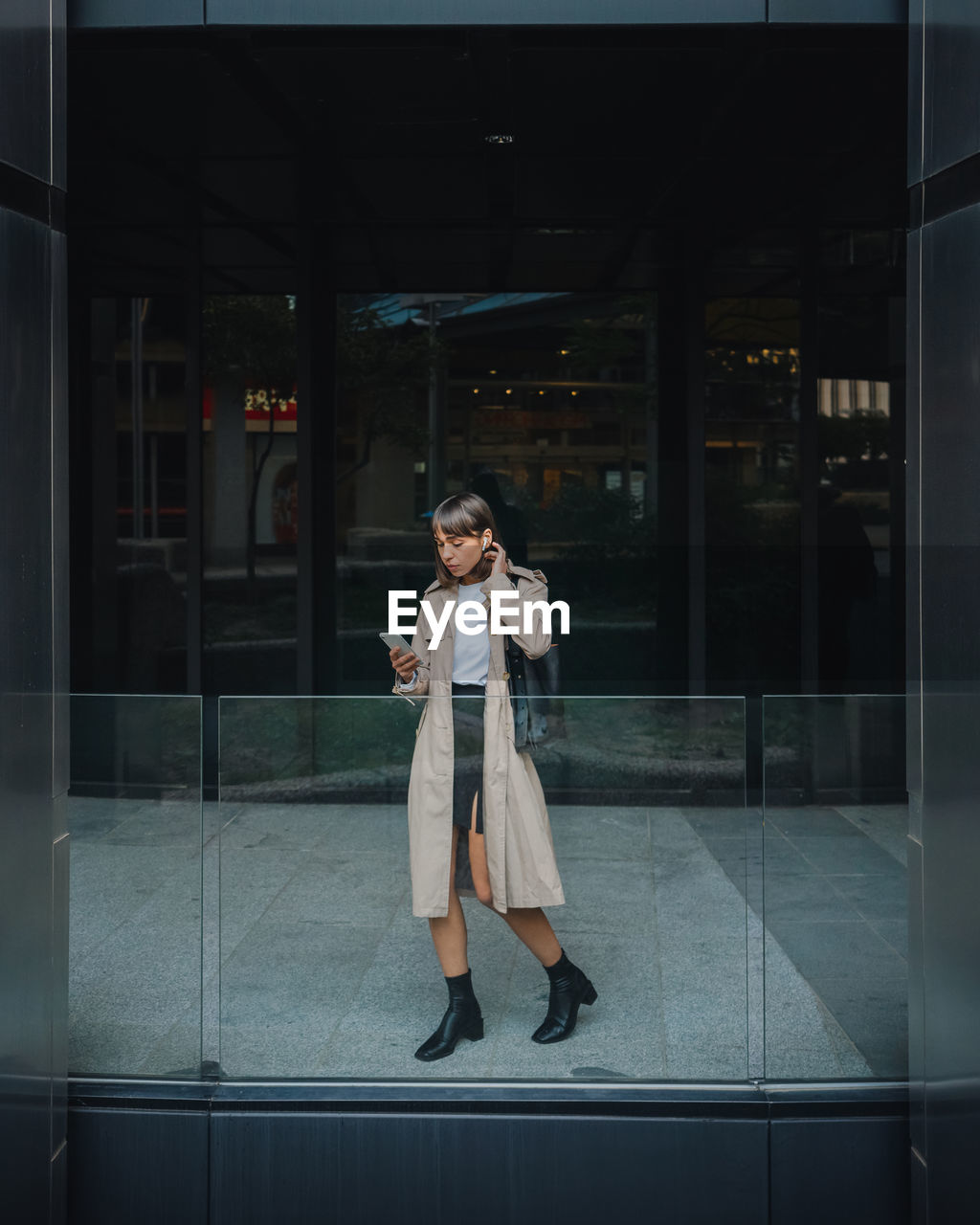 Full body of stylish female browsing cellphone while walking near contemporary building with glass fence on street of city