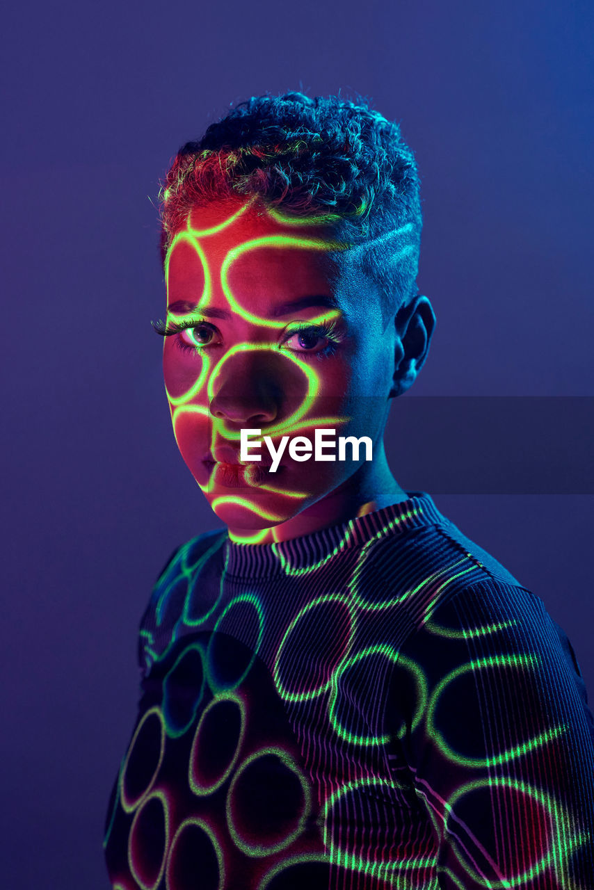 Charming african american female with short hair and in shadow projector light on face looking at camera on purple background in studio