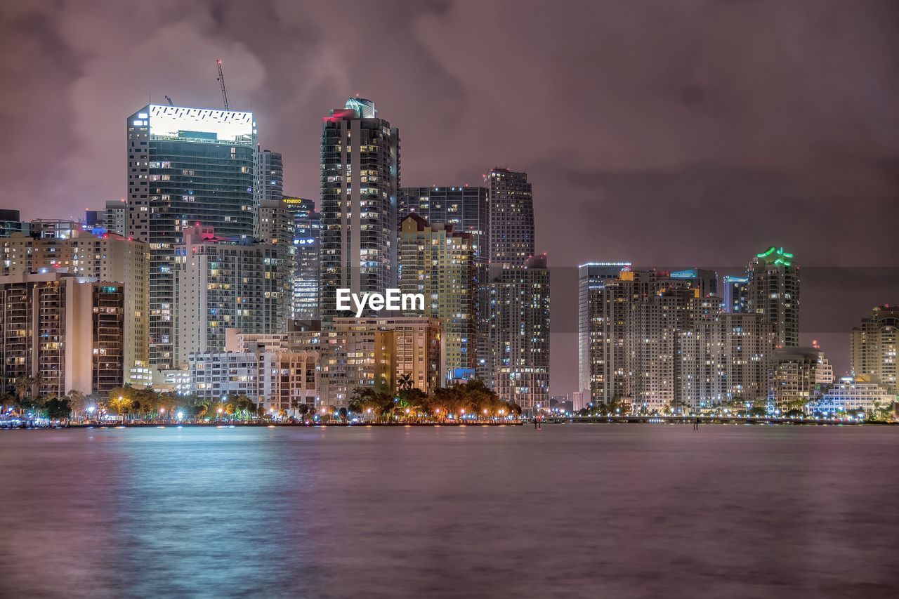 Sea and illuminated buildings against cloudy sky