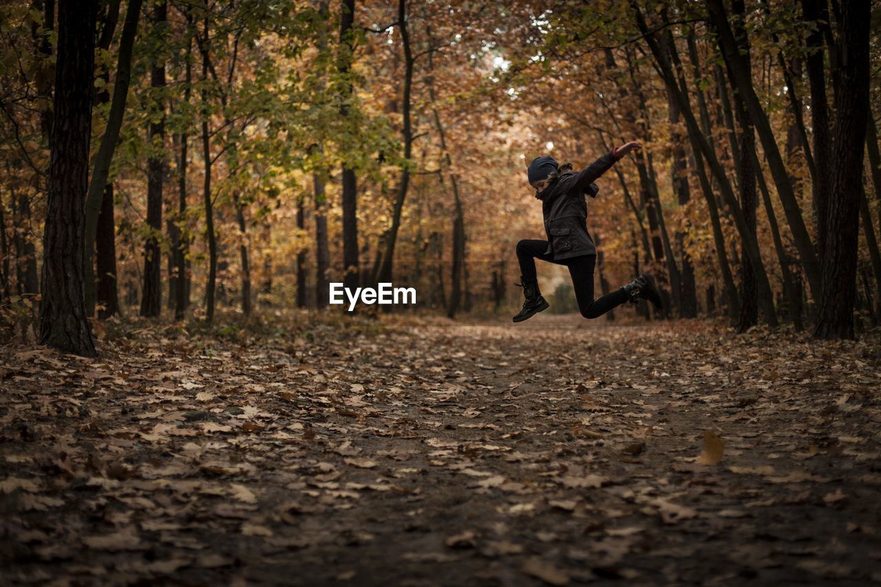 Side view of girl jumping on dirt road amidst trees in forest