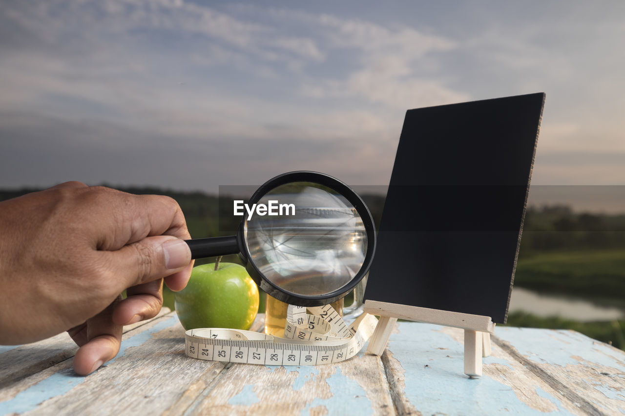 Cropped hand holding magnifying glass at table against sky