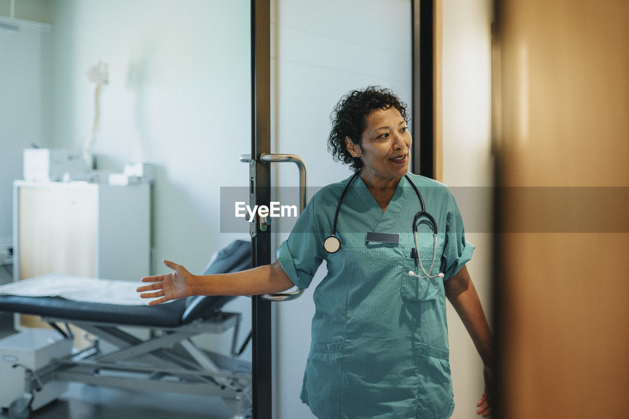 Female doctor wearing scrubs gesturing at doorway in hospital