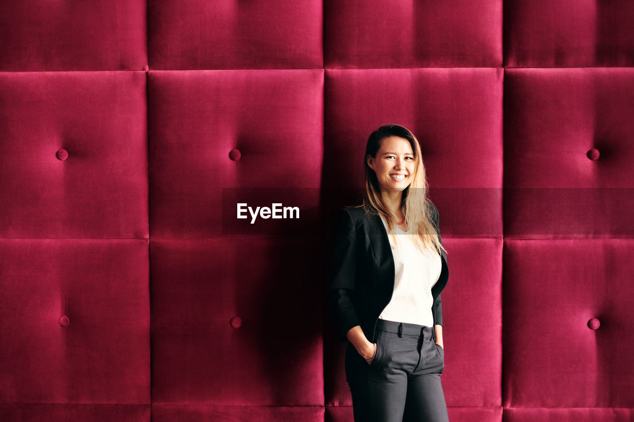 PORTRAIT OF SMILING YOUNG WOMAN AGAINST WALL