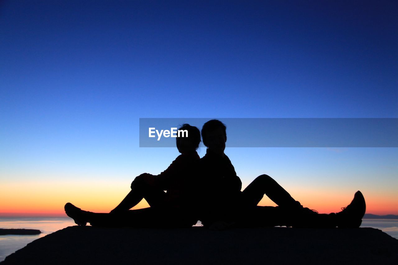 Silhouette couple sitting at beach against clear sky during sunset