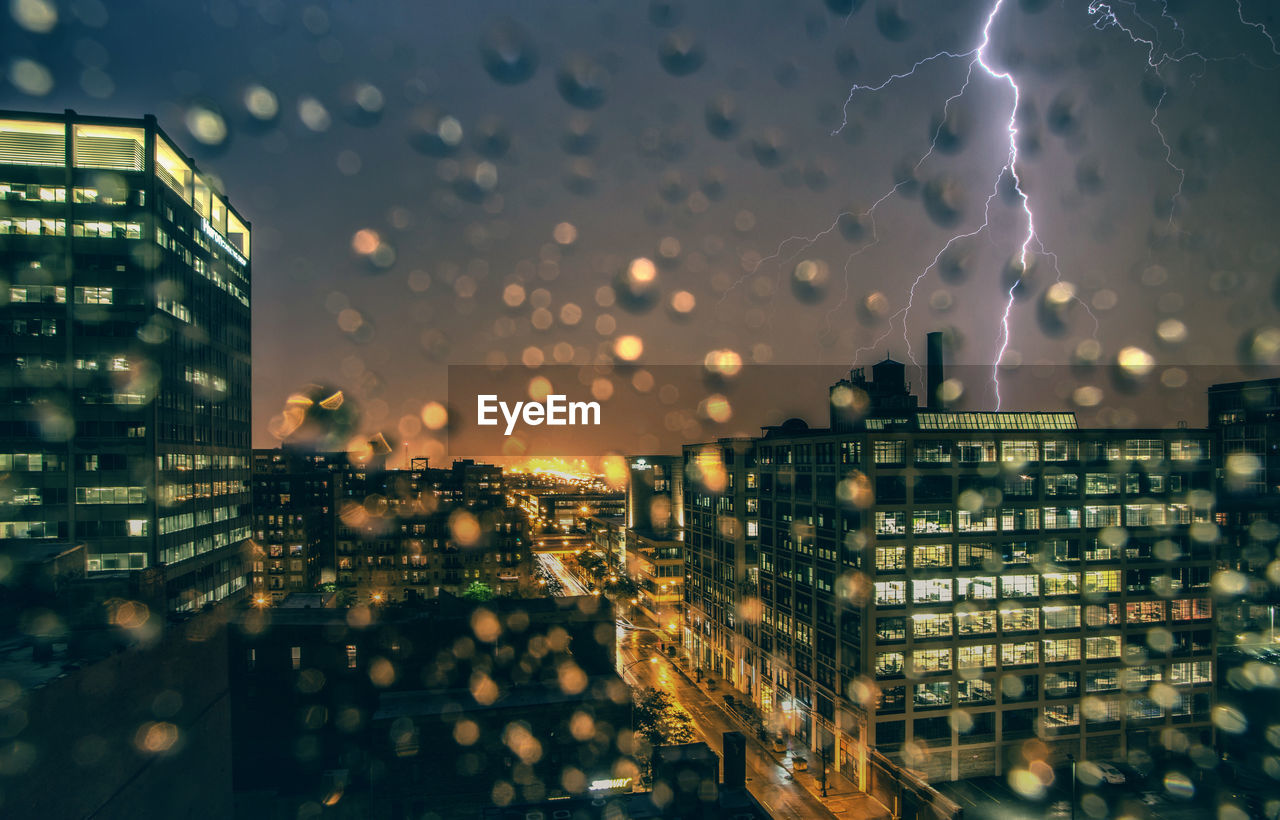 Illuminated buildings seen through window in city at night during thunderstorm