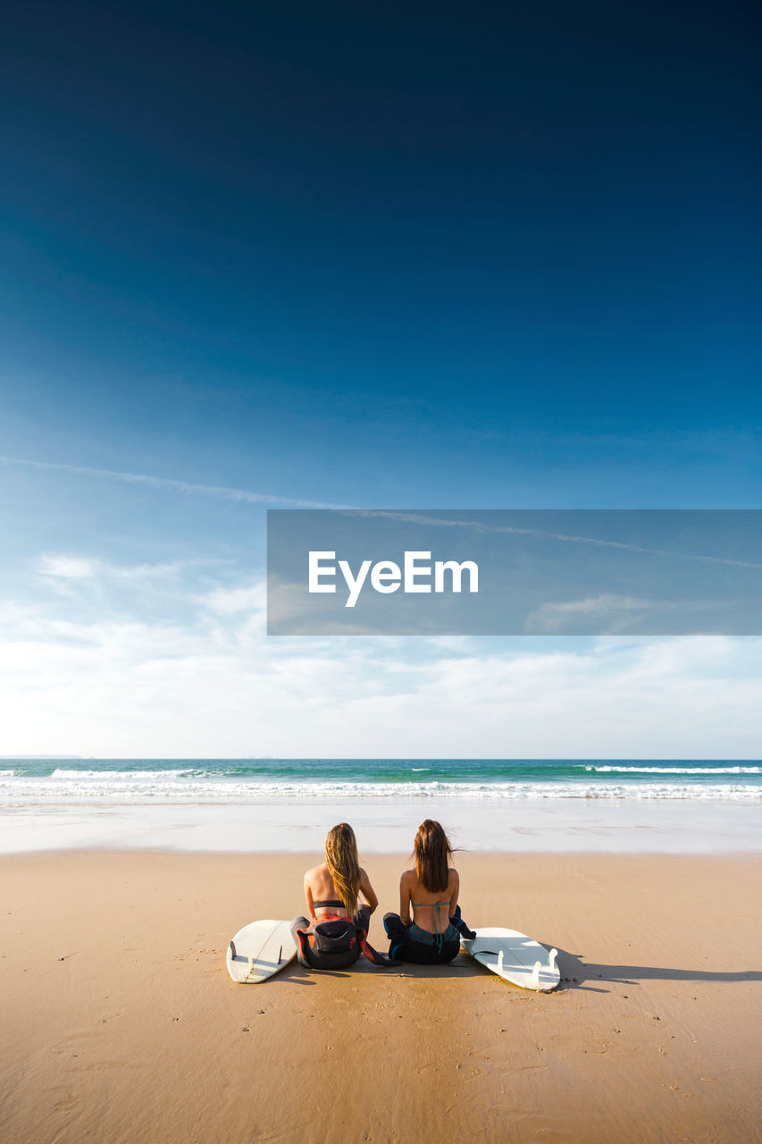 Rear view of female friends sitting at beach against blue sky