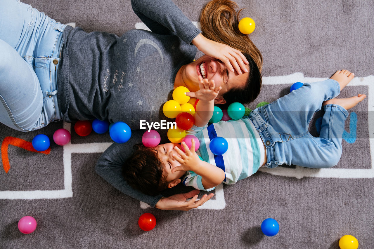 From above delighted woman and little boy screaming and throwing colorful balls up while lying on nursery floor together