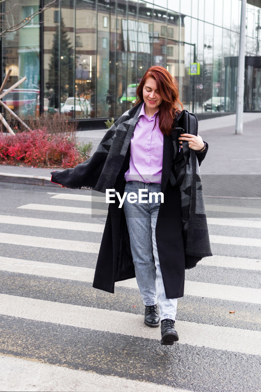city, one person, women, adult, architecture, city life, full length, street, smiling, young adult, bag, road, happiness, walking, crosswalk, transportation, clothing, casual clothing, city street, communication, lifestyles, hairstyle, coat, crossing, road marking, long hair, zebra crossing, building exterior, emotion, standing, technology, mobile phone, fashion, day, cheerful, outdoors, business, female, motion, on the move, built structure, shopping bag, redhead, wireless technology, telephone, looking, jacket, portrait, smartphone, footwear, symbol, front view, holding, sign, leisure activity, using phone, smile, outerwear, person, brown hair, teeth, shopping, marking, businesswoman