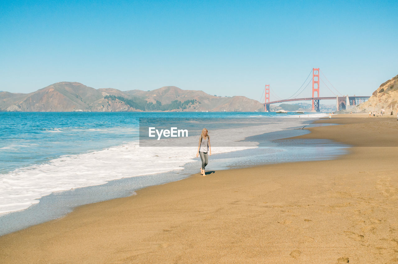 Woman walking on shore with golden gate bridge against clear sky
