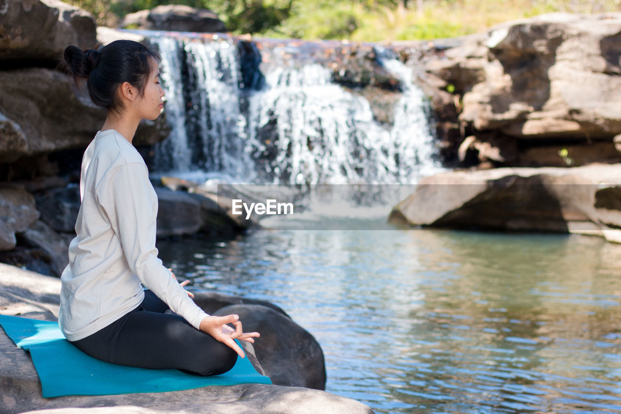 Side view of woman doing lotus position yoga while sitting on rocks against waterfall
