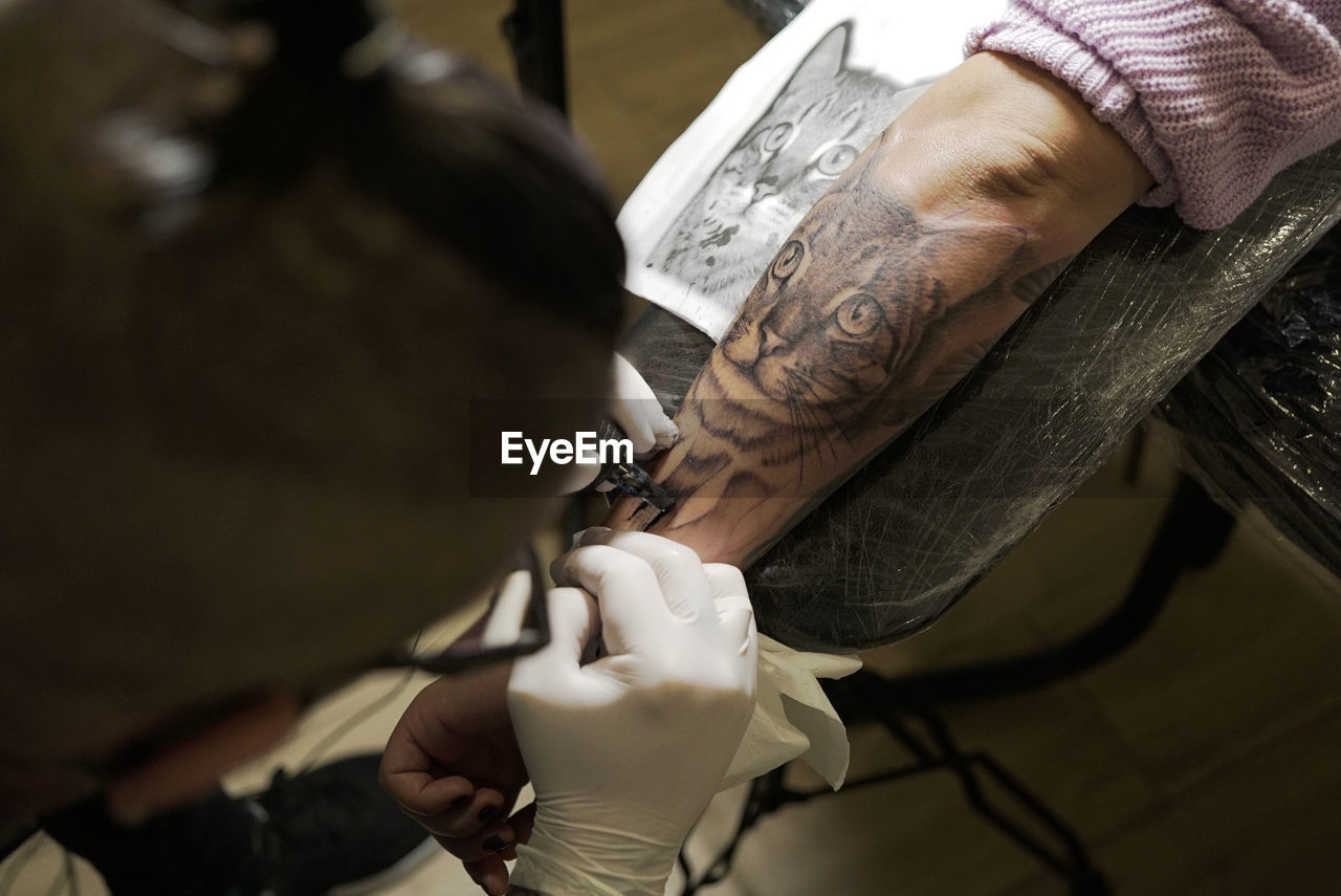 From above of unrecognizable male tattooist using machine and making tattoo on arm of crop client in salon