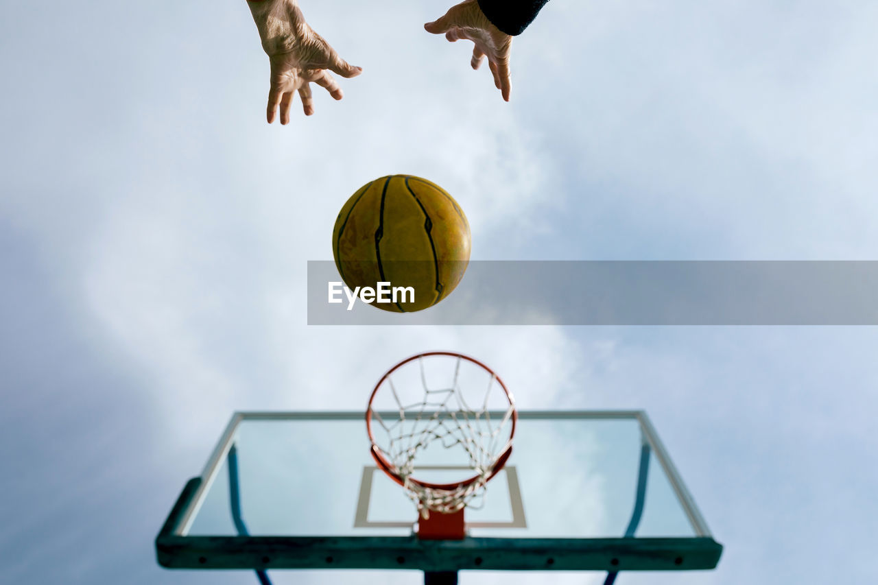 From below of crop anonymous person throwing basketball ball into hoop while playing game on public sports ground against blue sky