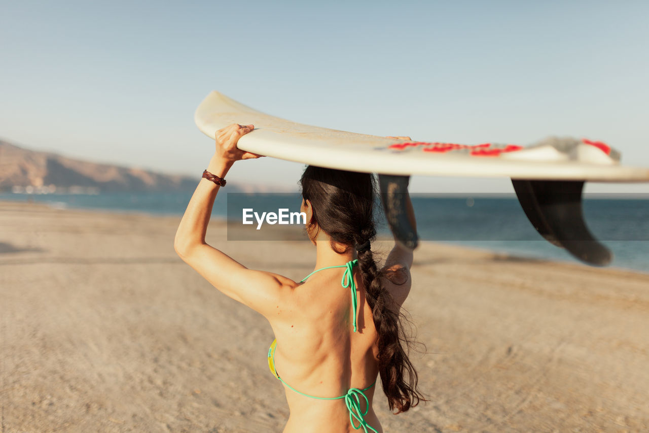 Rear view of woman carrying surfboard while walking at beach