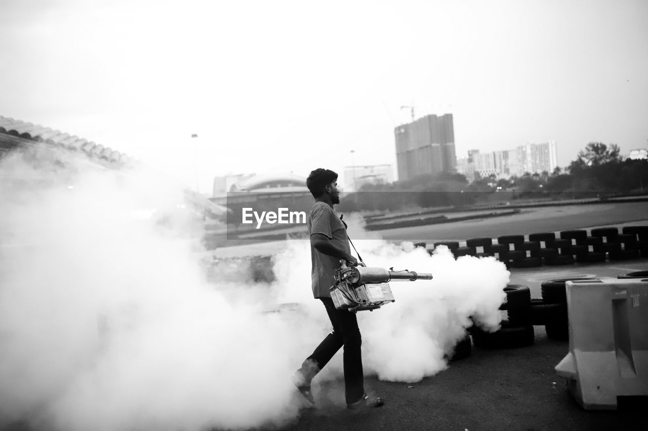 Full length of man holding mosquito fogging machinery against sky in city