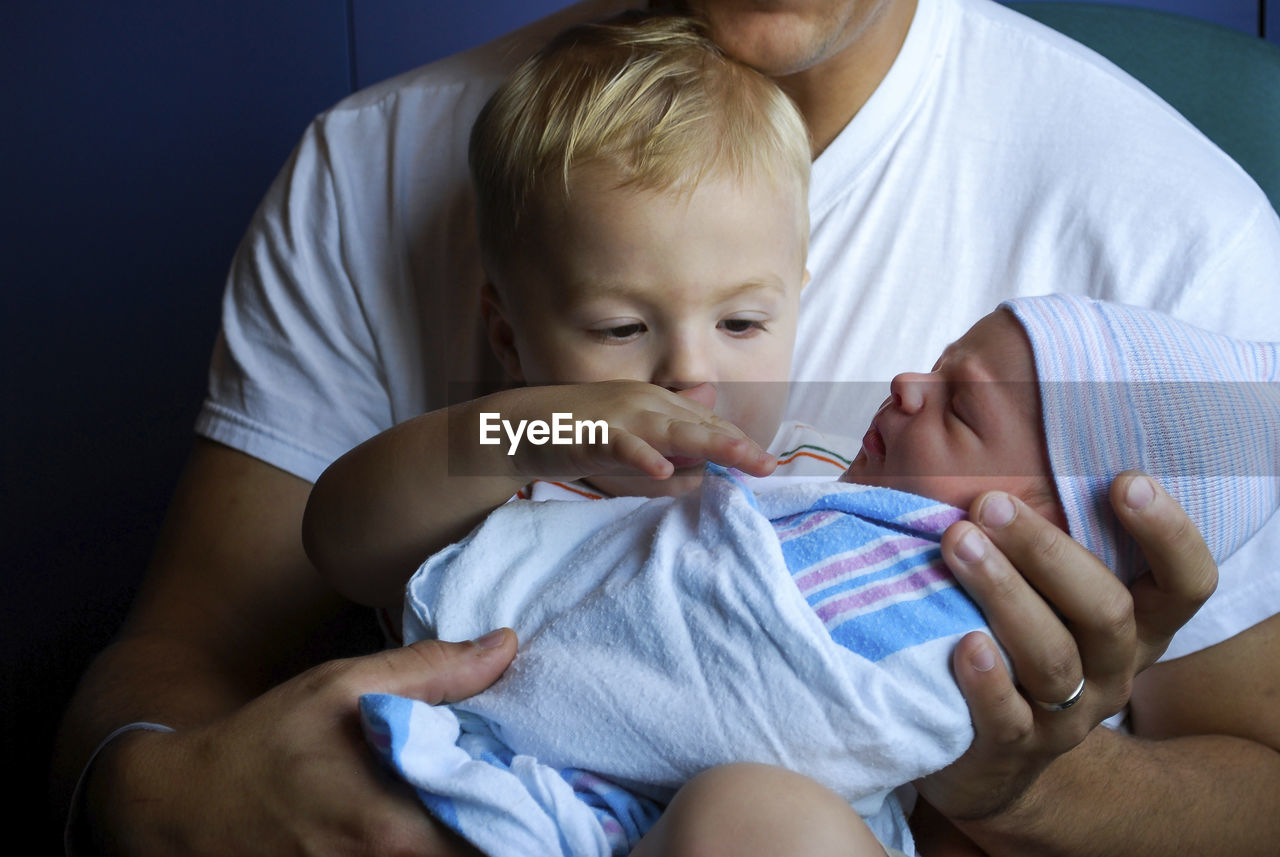 Toddler touching newborn brother wrapped in blanket being carried by father