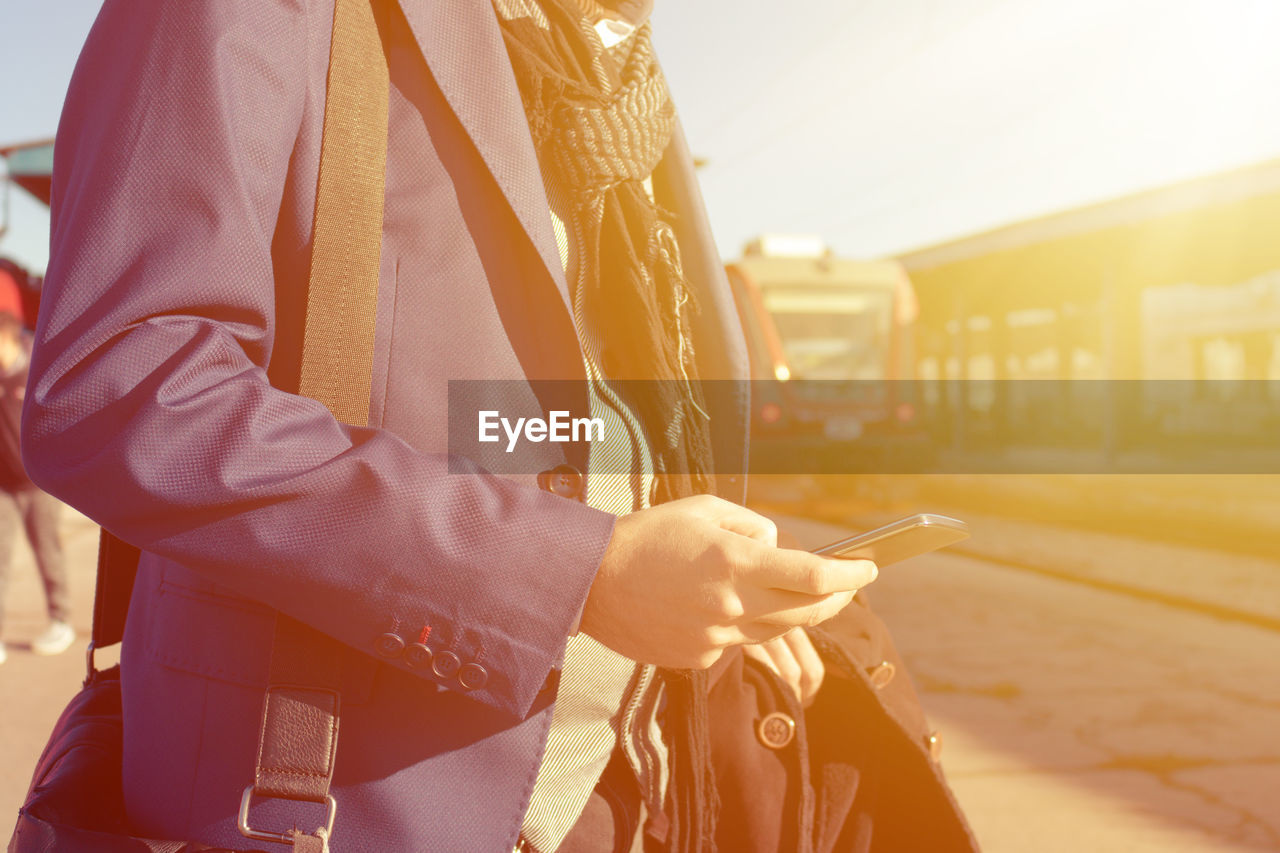 Midsection of man holding mobile phone while standing at railroad station platform