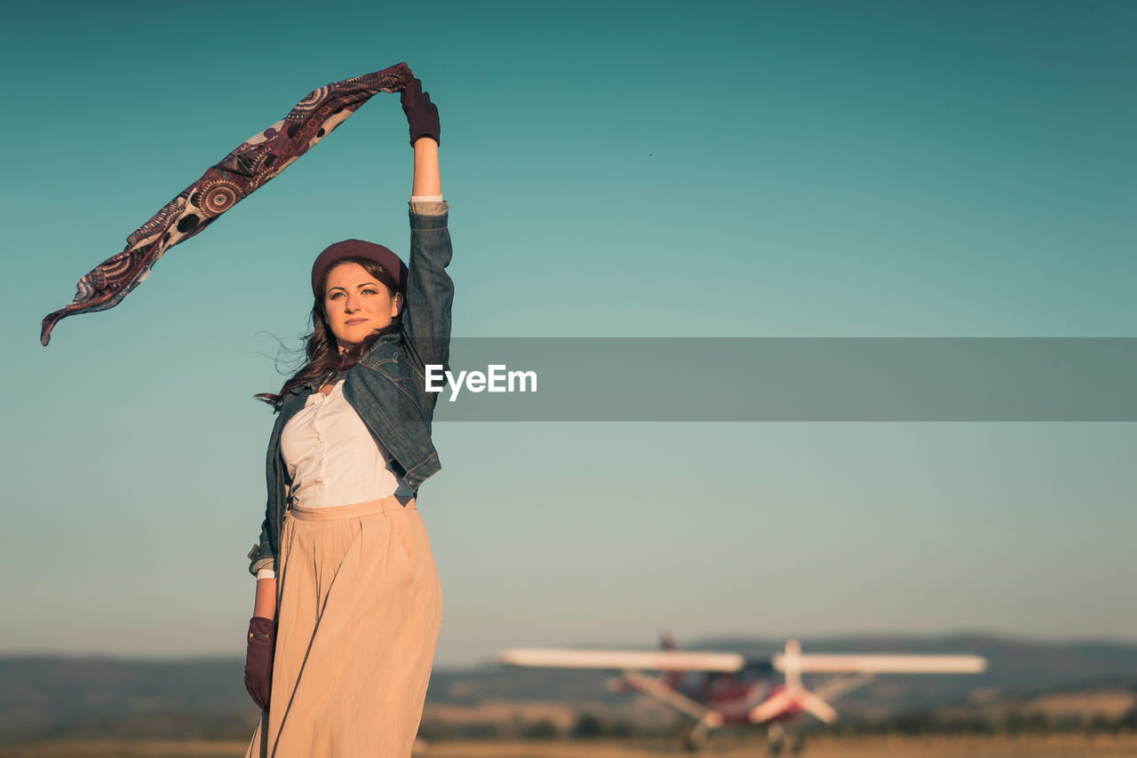 PORTRAIT OF YOUNG WOMAN STANDING IN FRONT OF TRADITIONAL WINDMILL AGAINST CLEAR SKY