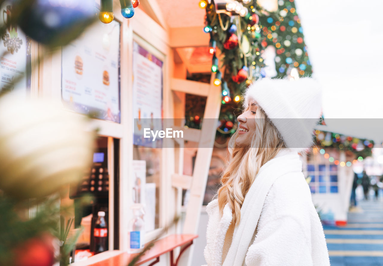 Young happy woman with curly hair in white knitted hat on shopping at christmas fair market 