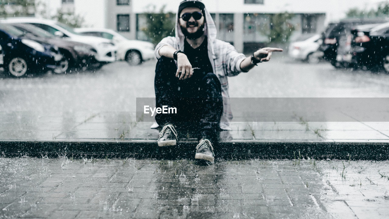 Portrait of young man gesturing while sitting on footpath during rain