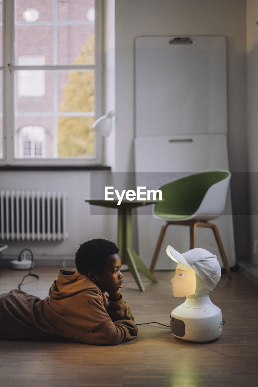 Boy talking with illuminated ai robot while lying on floor in innovation lab